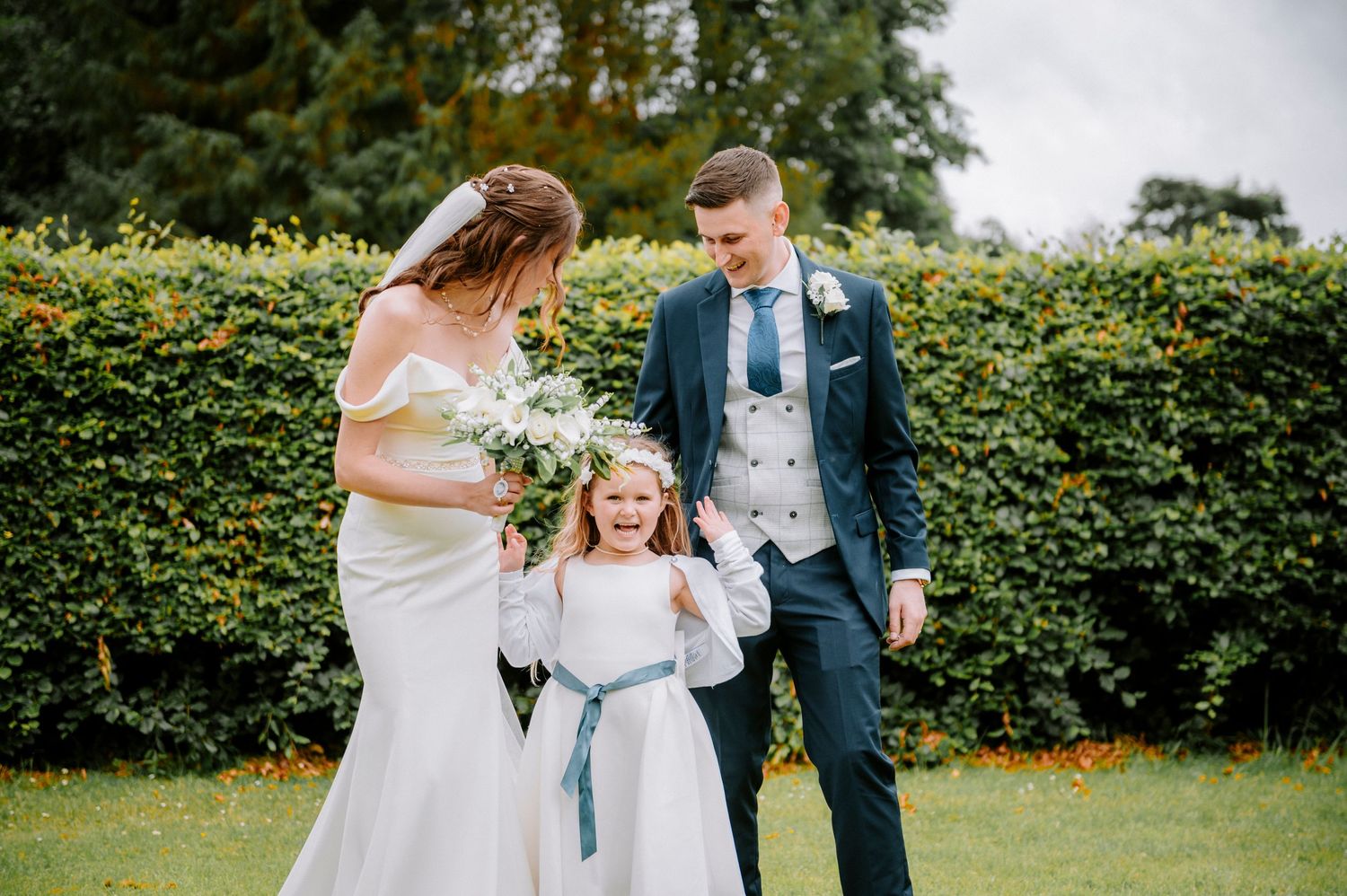 Wedding party poses in front of lush green hedges on a sunny day.