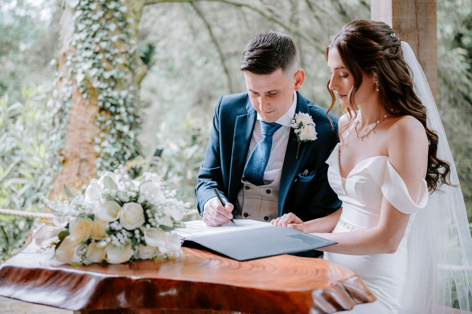 Wedding ceremony signing documents in a rustic outdoor venue with white floral arrangements.
