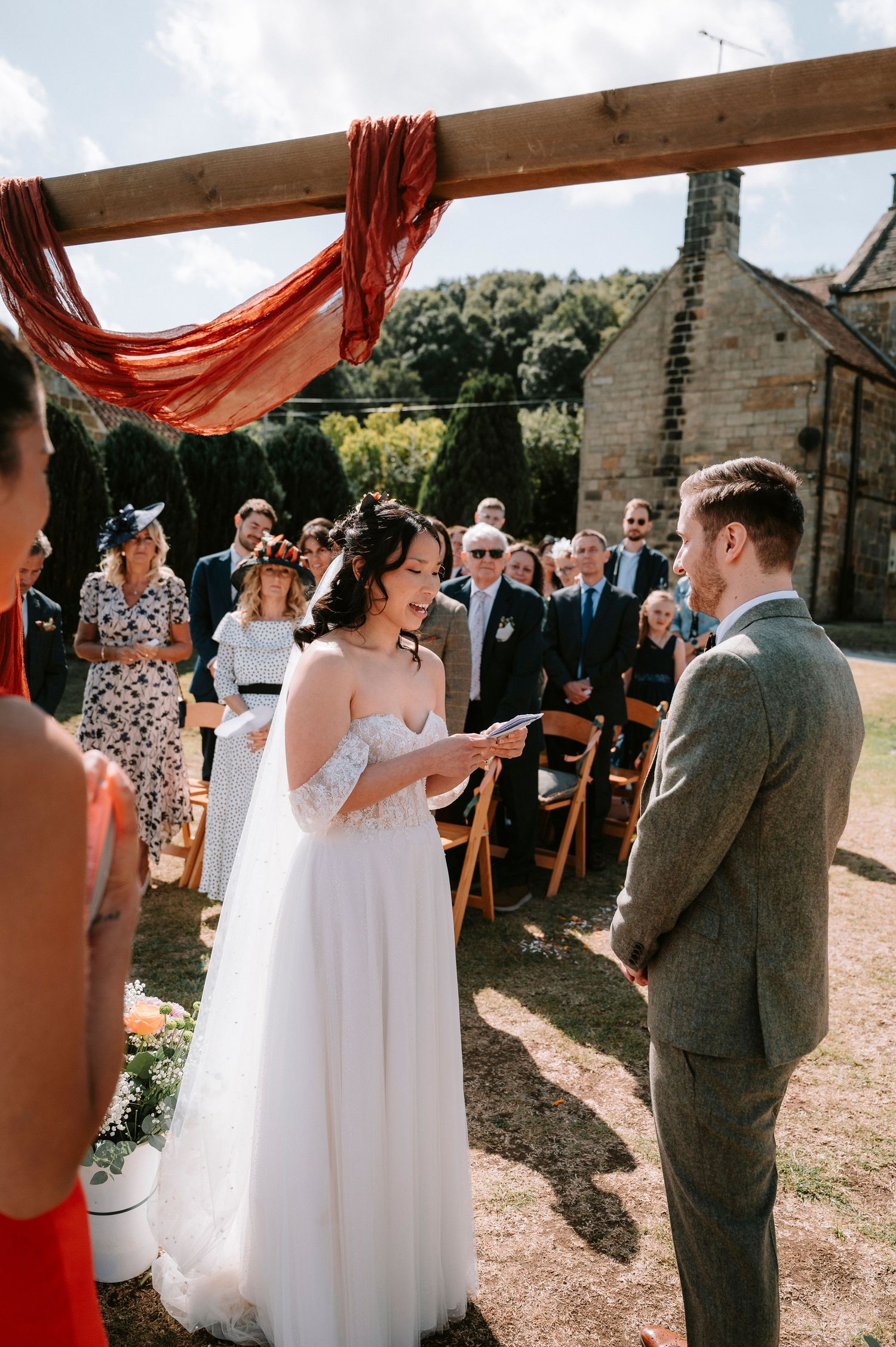 Outdoor wedding ceremony with red fabric decor and stone building backdrop.