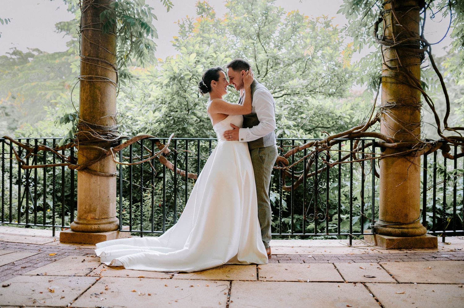 A romantic wedding couple embraces under ornate stone pillars and iron railings.