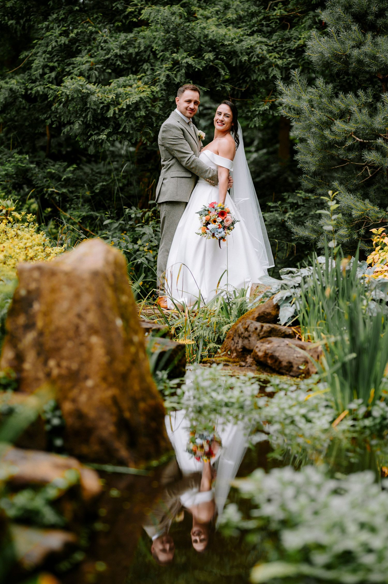 Garden wedding portraits with reflection in water feature among flowers and rocks.