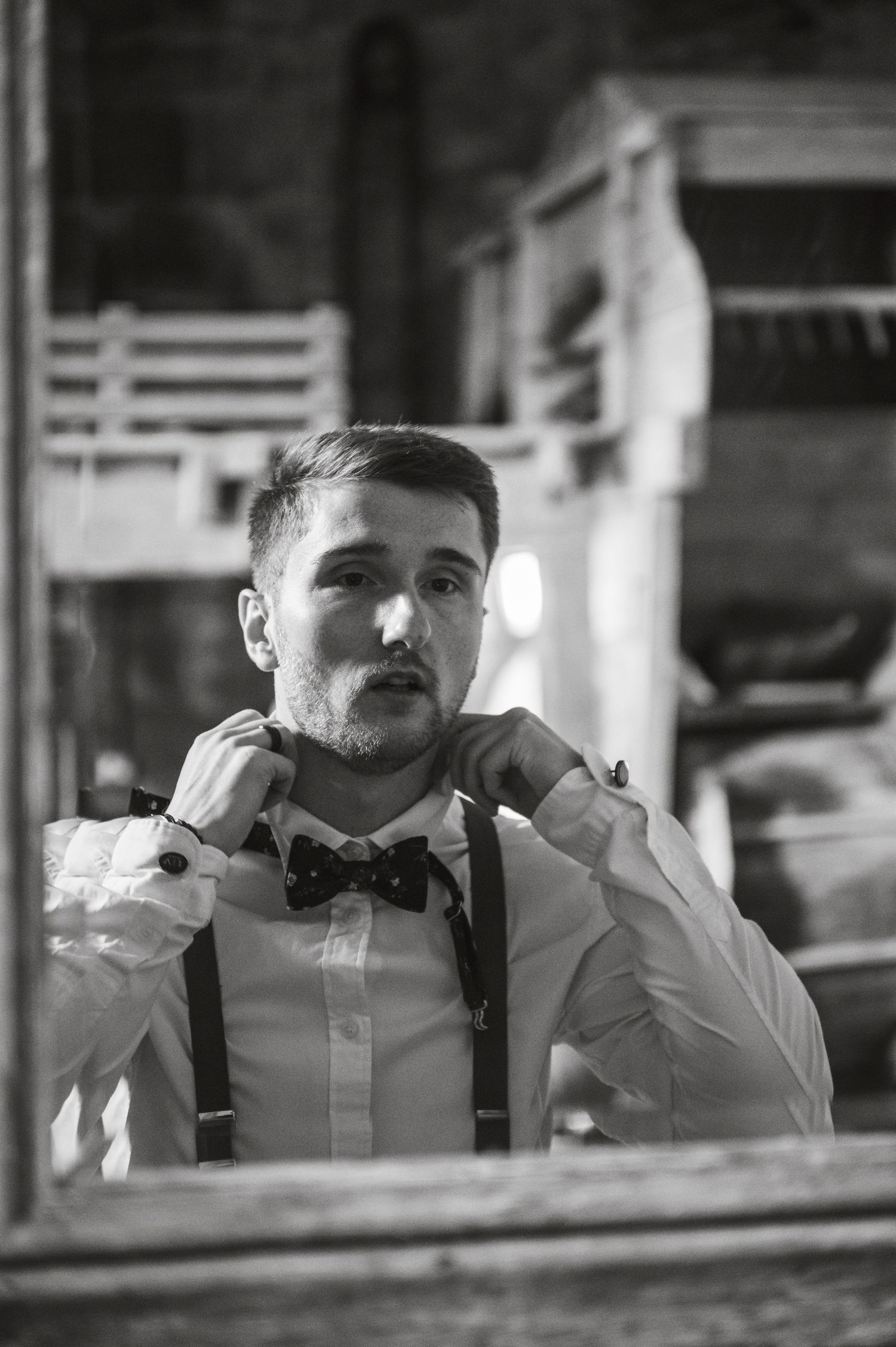 A black and white reflection of someone adjusting their bowtie and suspenders in a mirror.