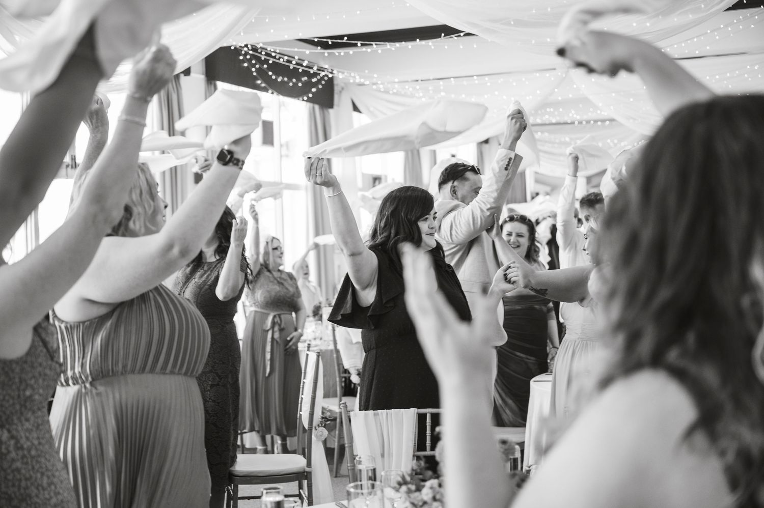 Wedding reception guests dancing and celebrating with raised arms in black and white.