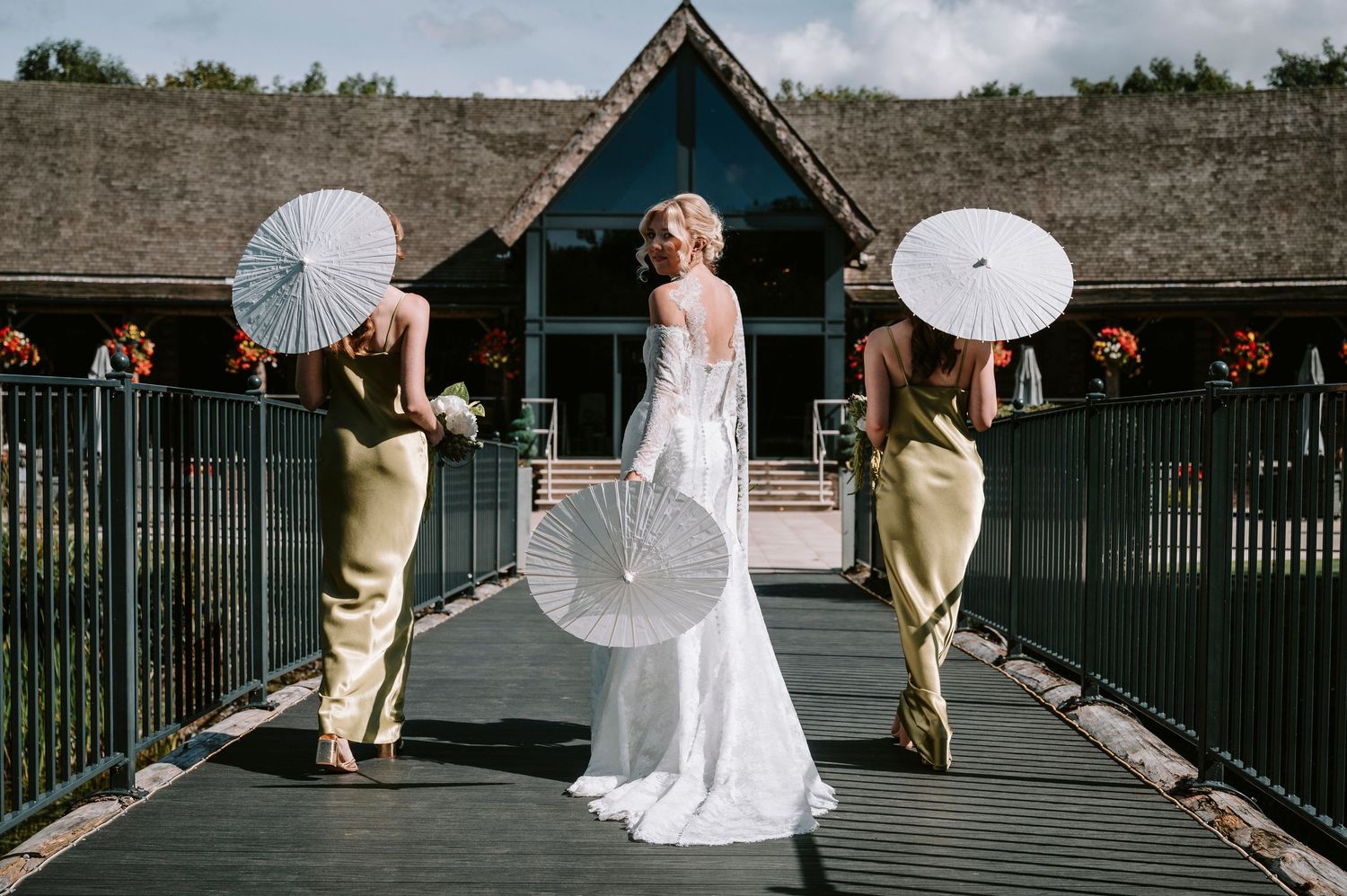 A wedding party poses with parasols on a wooden boardwalk surrounded by rustic buildings on a cloudy day.