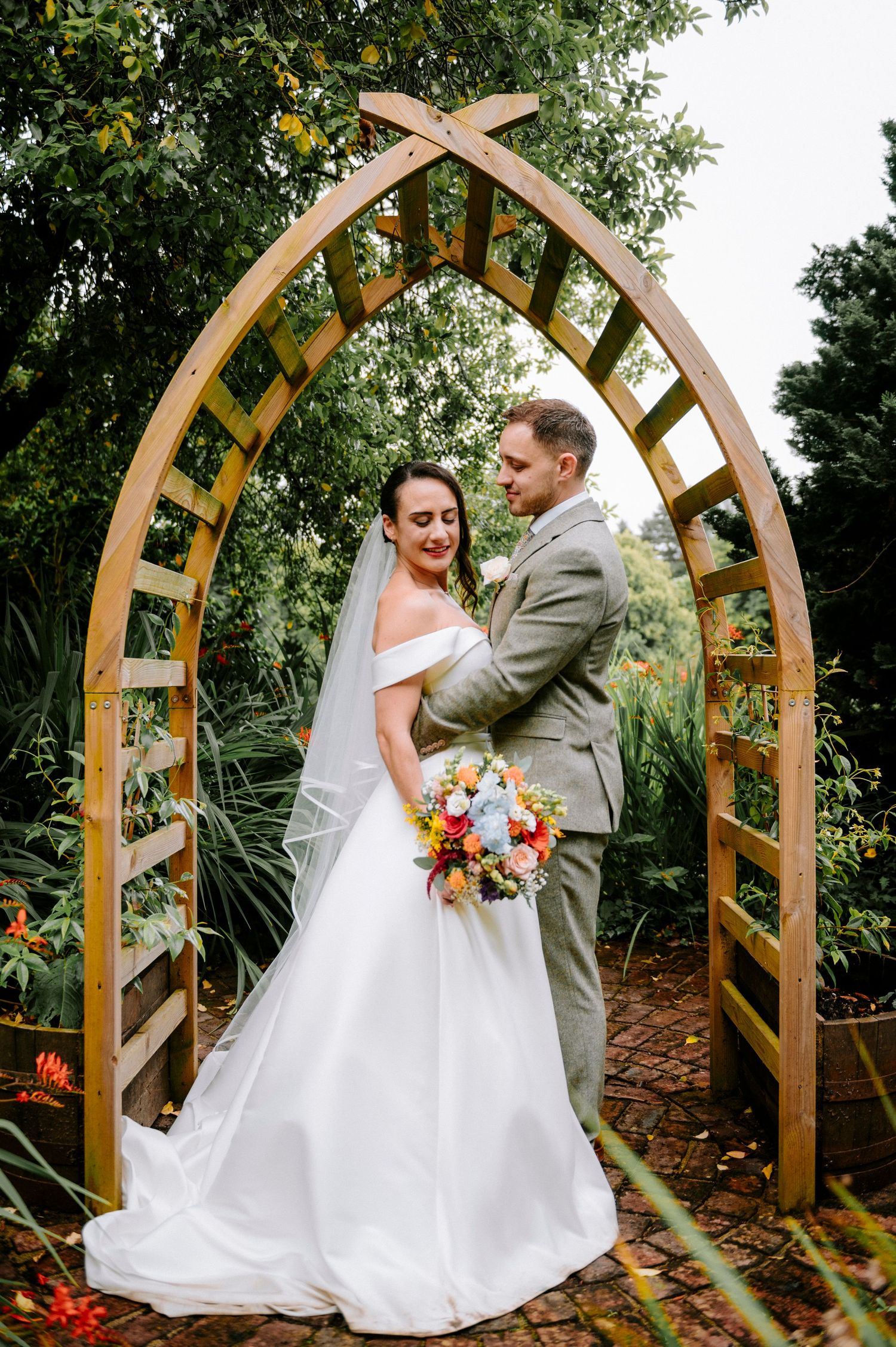 Wedding couple posing under wooden garden arbor with natural landscaping.