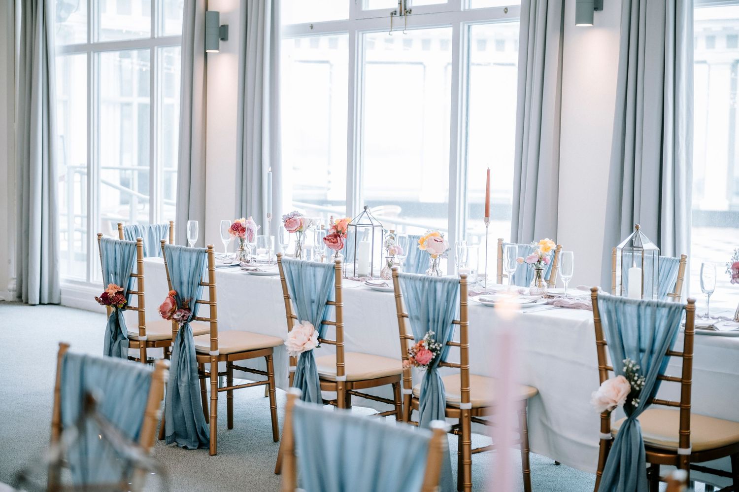 A bright reception venue with blue chairs and elegant table settings by large windows.