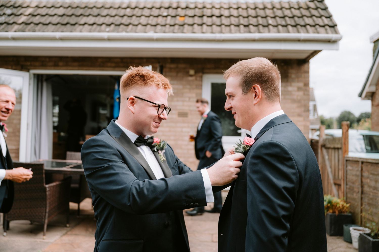 Groomsmen helping each other with formal wear preparations outdoors.