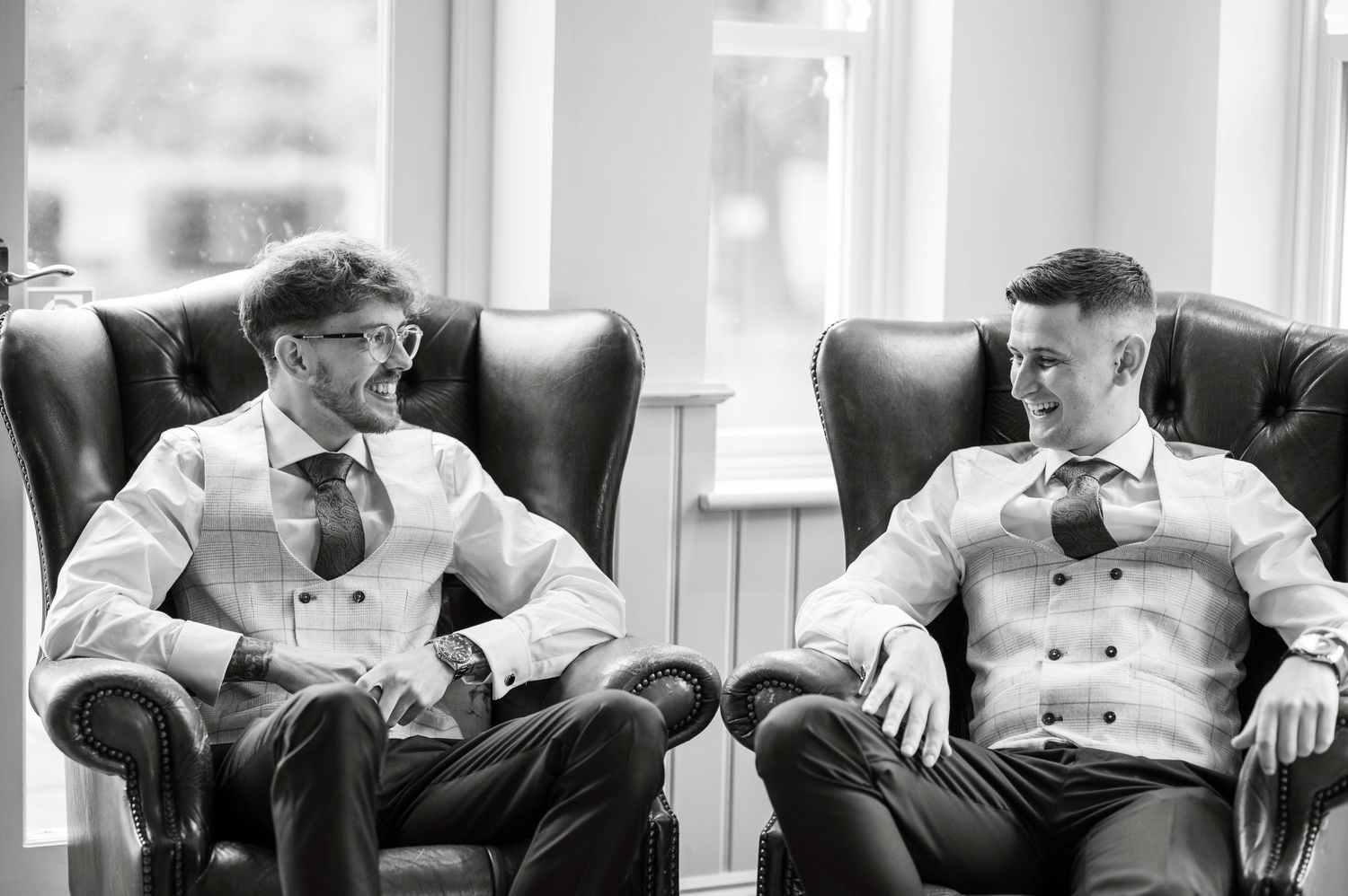 Two people in formal attire sharing a laugh while seated in leather armchairs.