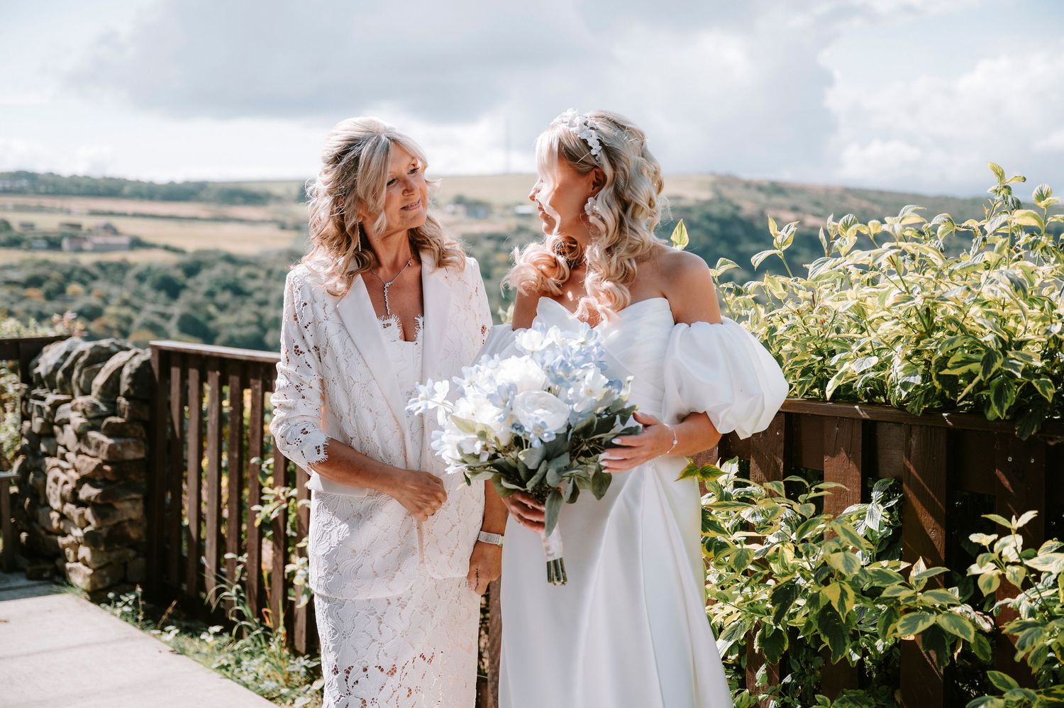 Two women in white dresses share a moment while holding bouquets on an outdoor terrace.