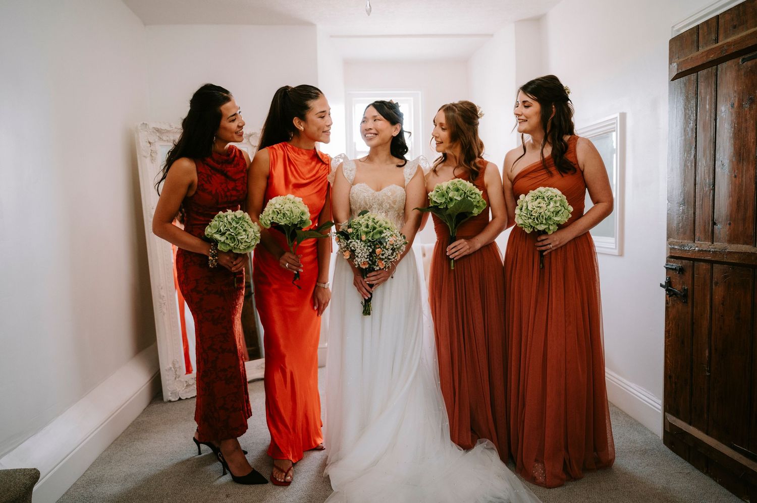 Wedding party members in burgundy and rust-colored dresses standing together holding bouquets.
