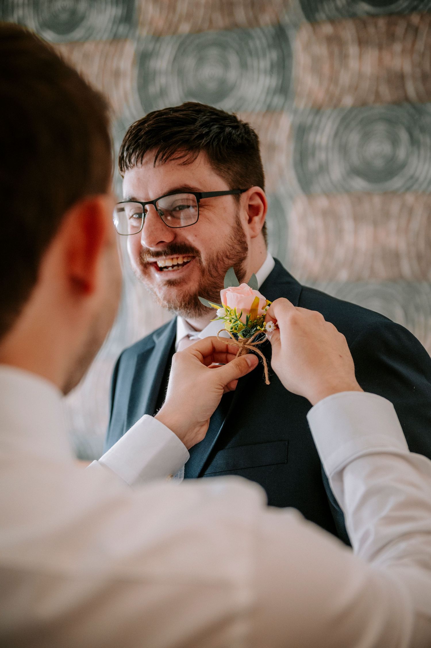 Groomsman helps adjust boutonniere on suit jacket during wedding preparations.