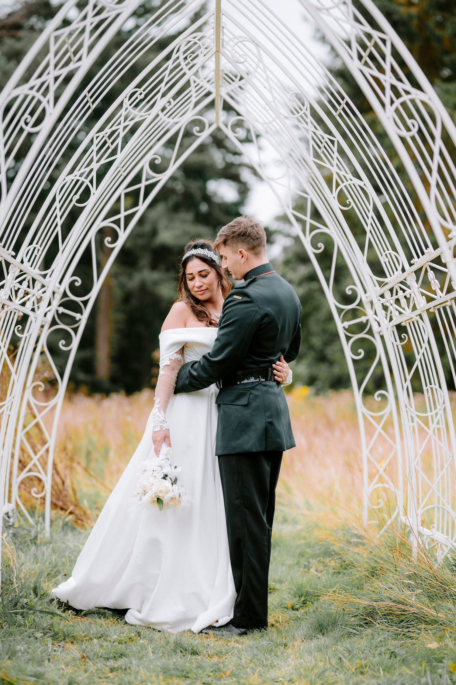 Newlyweds share an intimate moment beneath a decorative white metal archway in a garden setting.