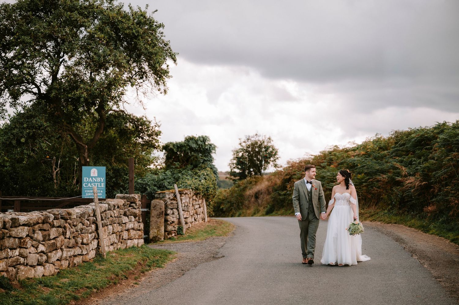 Wedding party walking along a stone wall path through countryside scenery.