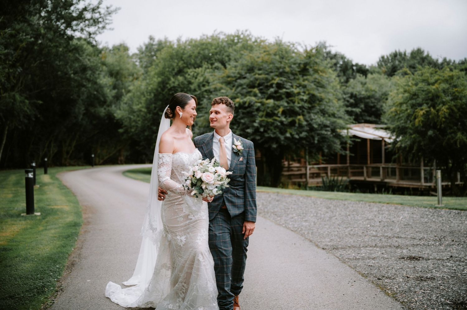 A wedding couple walks along a tree-lined gravel path outdoors.