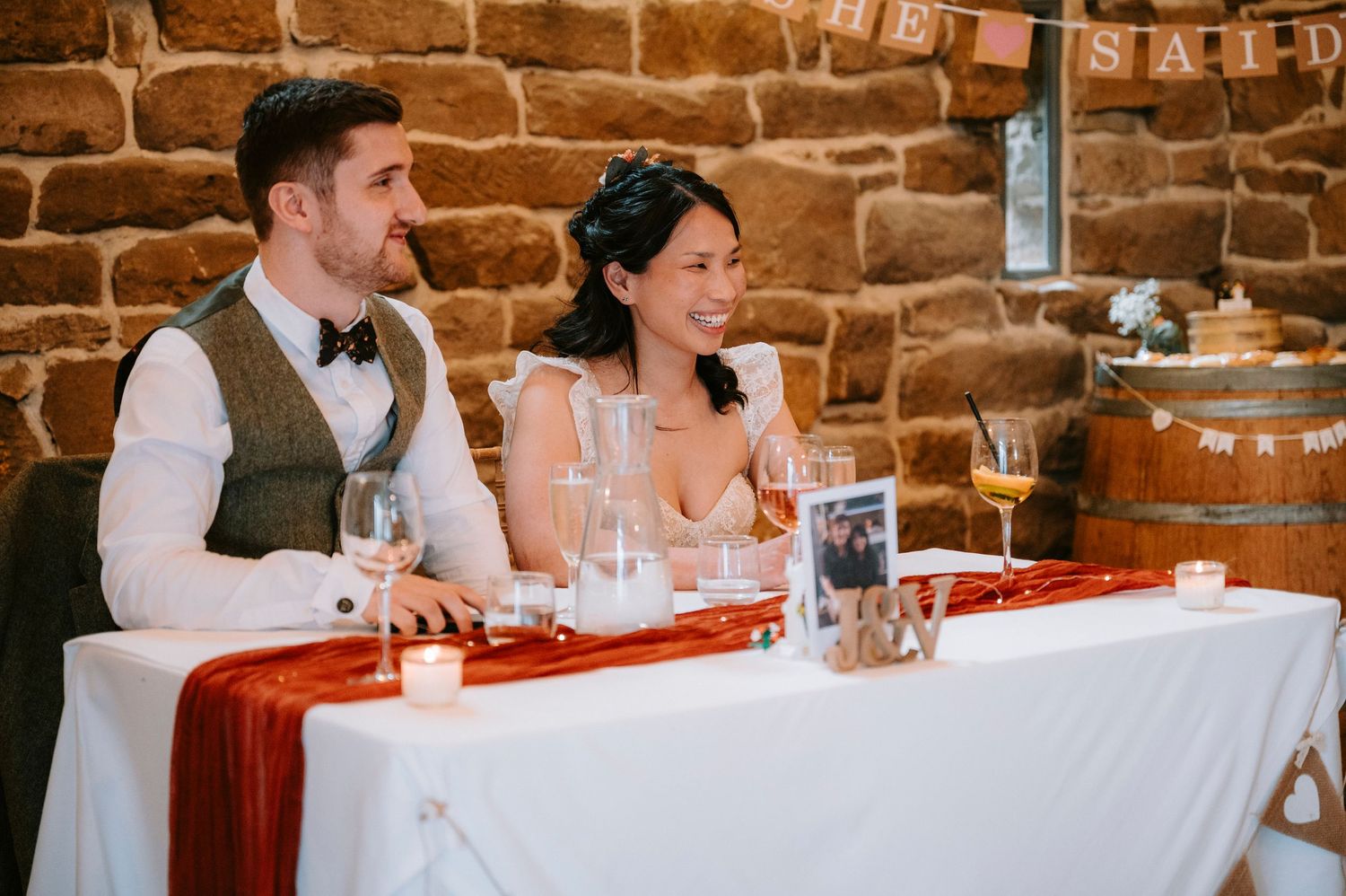 A couple sits at their wedding head table in a rustic brick-walled venue during their reception.