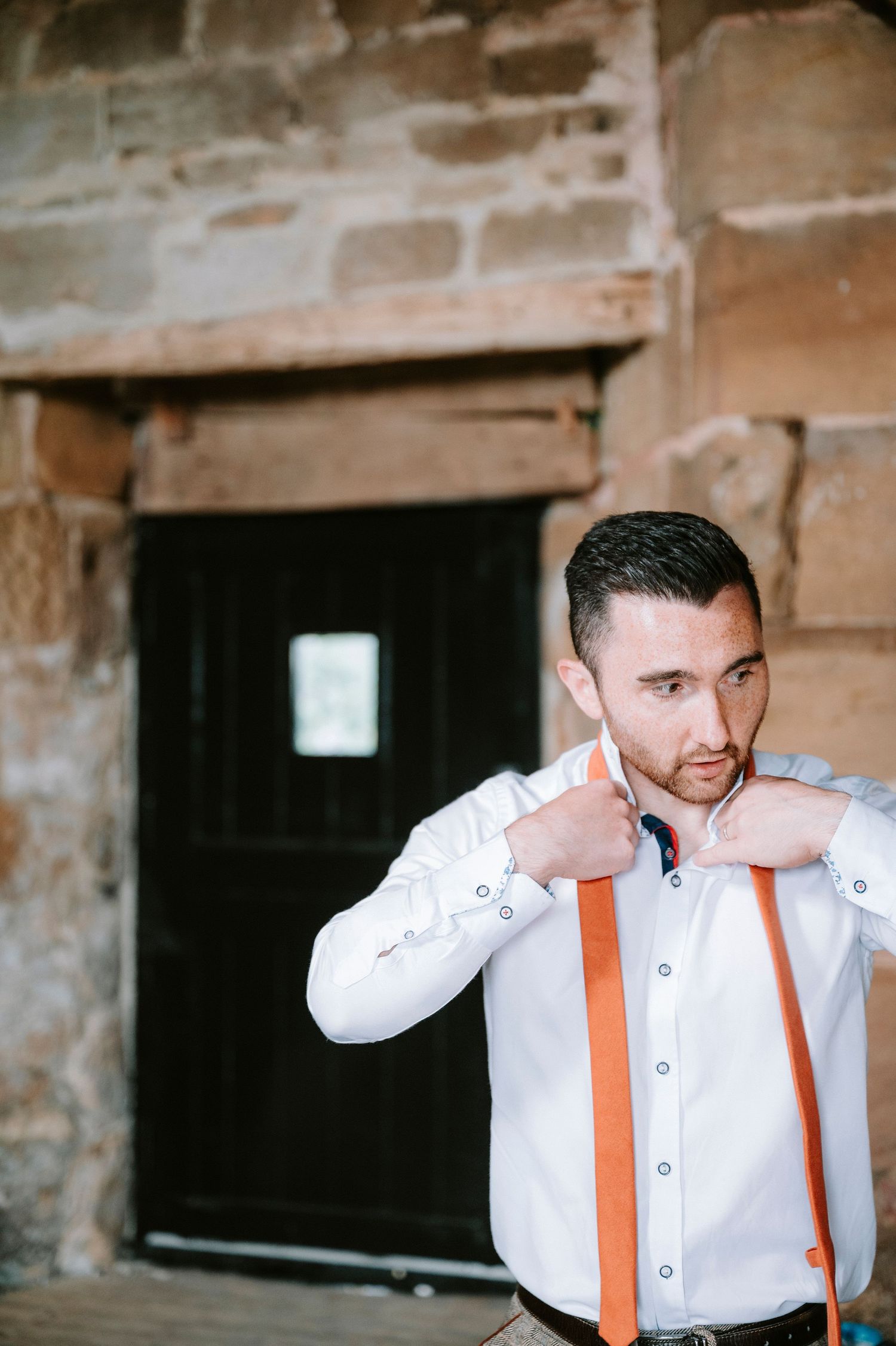 Person adjusting orange suspenders while wearing a white dress shirt against a stone wall background.