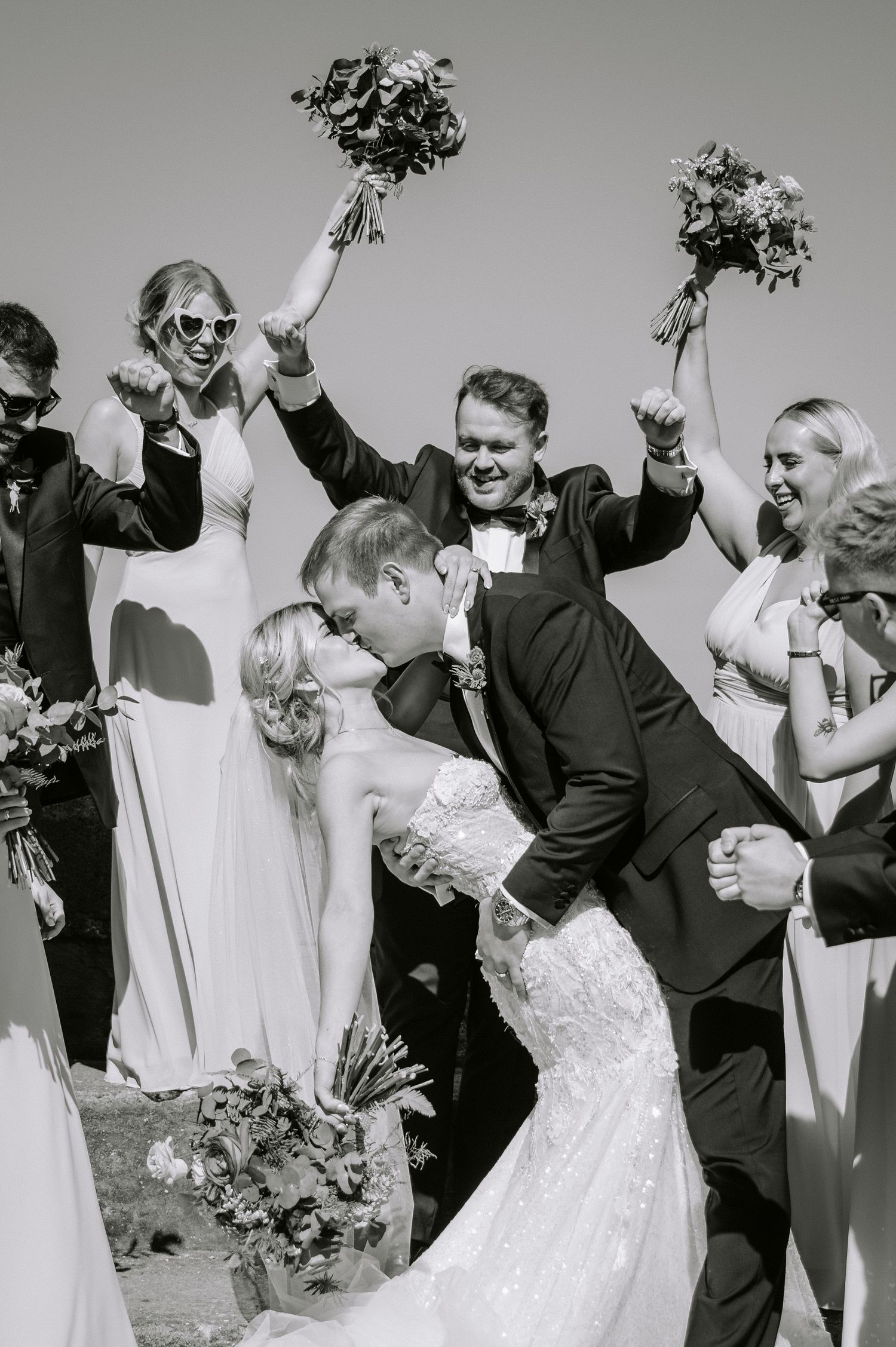 Wedding party celebrates with raised bouquets as newlyweds share romantic dip kiss in black and white photo.