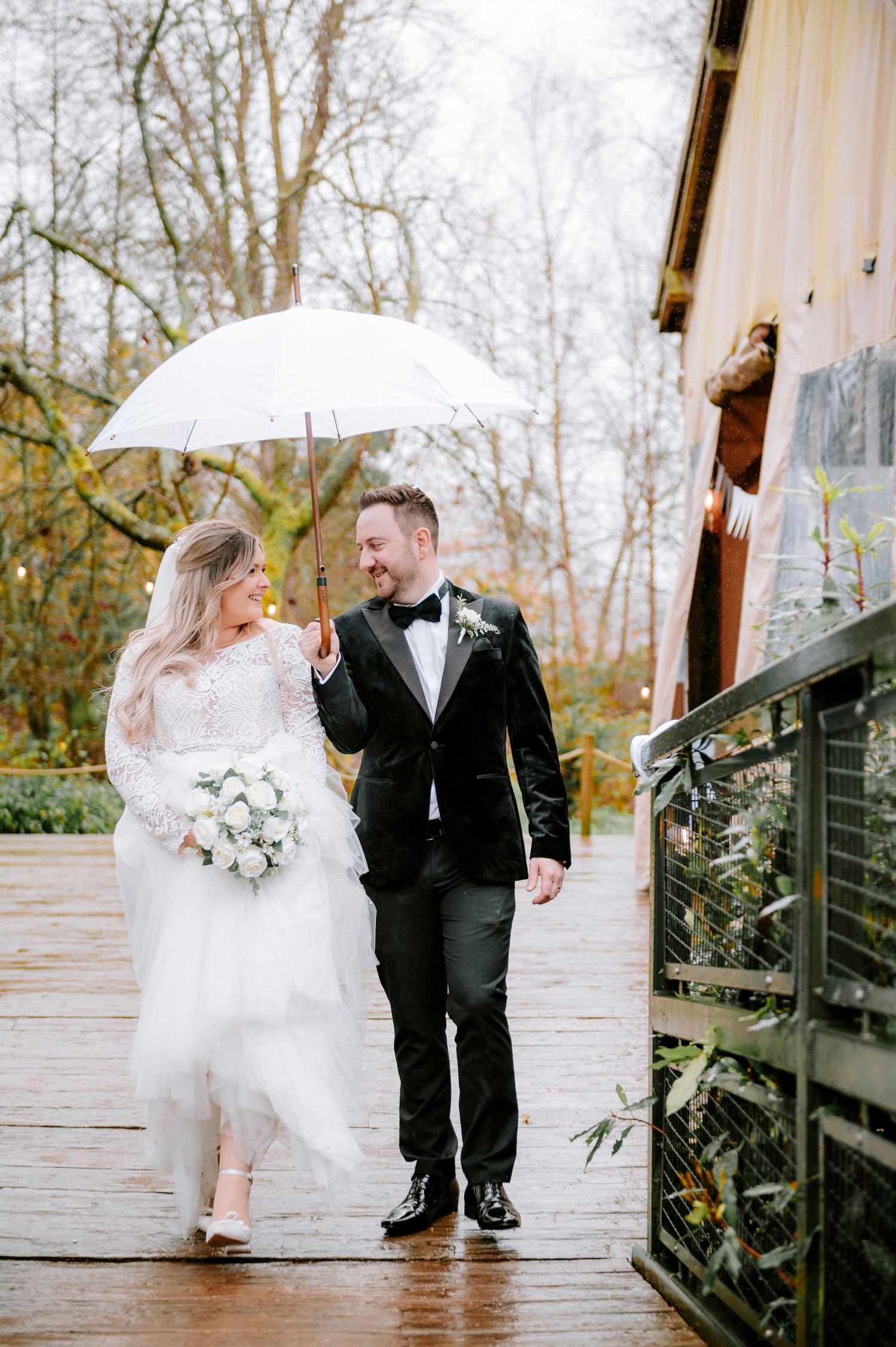 A couple walks together under a white umbrella on a rainy wedding day on a wooden deck.