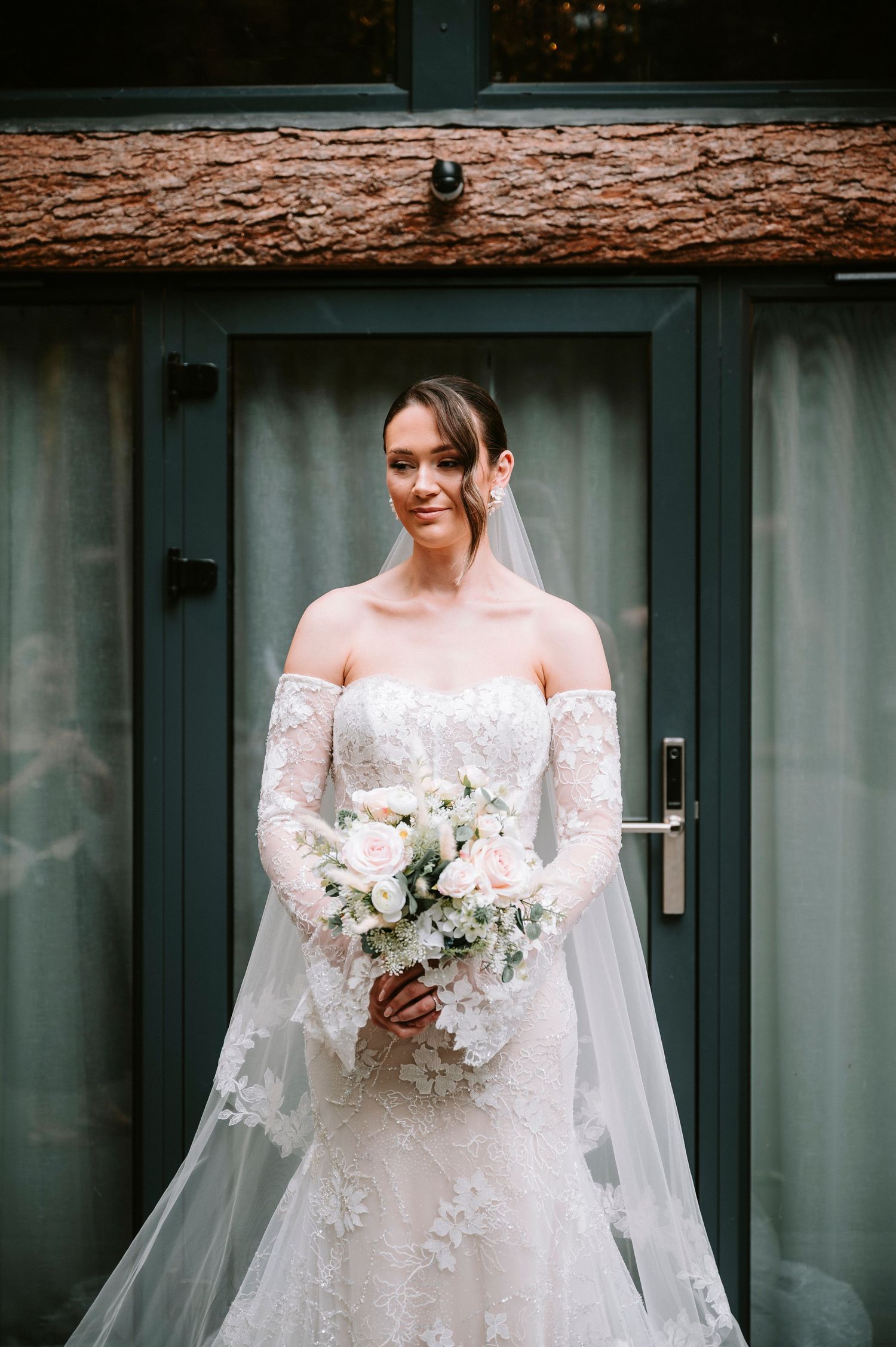 A bride in an elegant long-sleeved lace gown stands against a modern gray exterior wall.