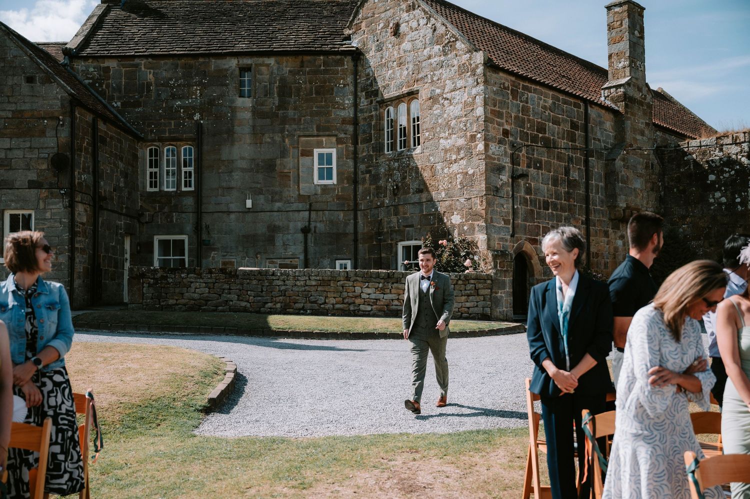 Wedding guests gather outside a rustic stone building on a sunny day.