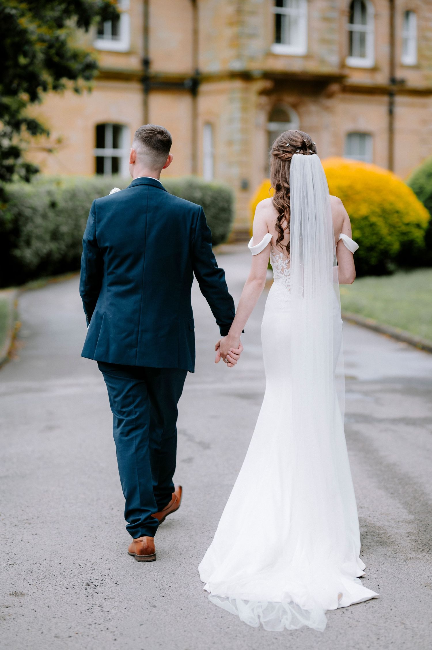 Newlyweds walking together on a path outside a stone building with flowing white dress and veil.