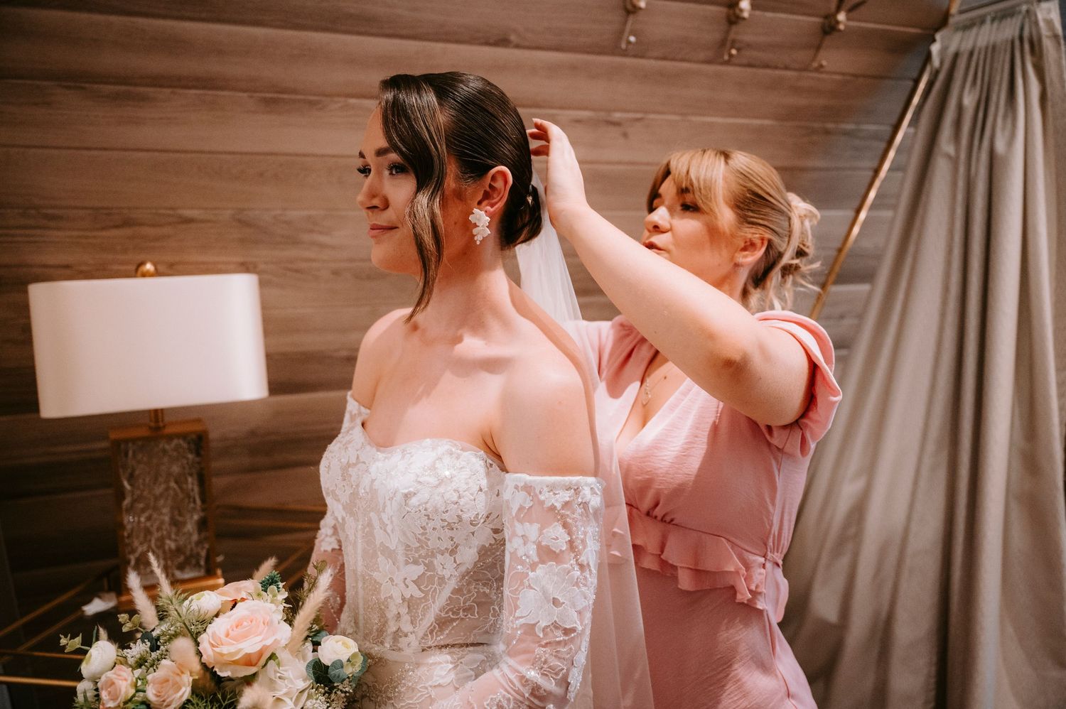 A bride in a lace wedding dress holds a bouquet of white and pink flowers against a rustic wooden wall.