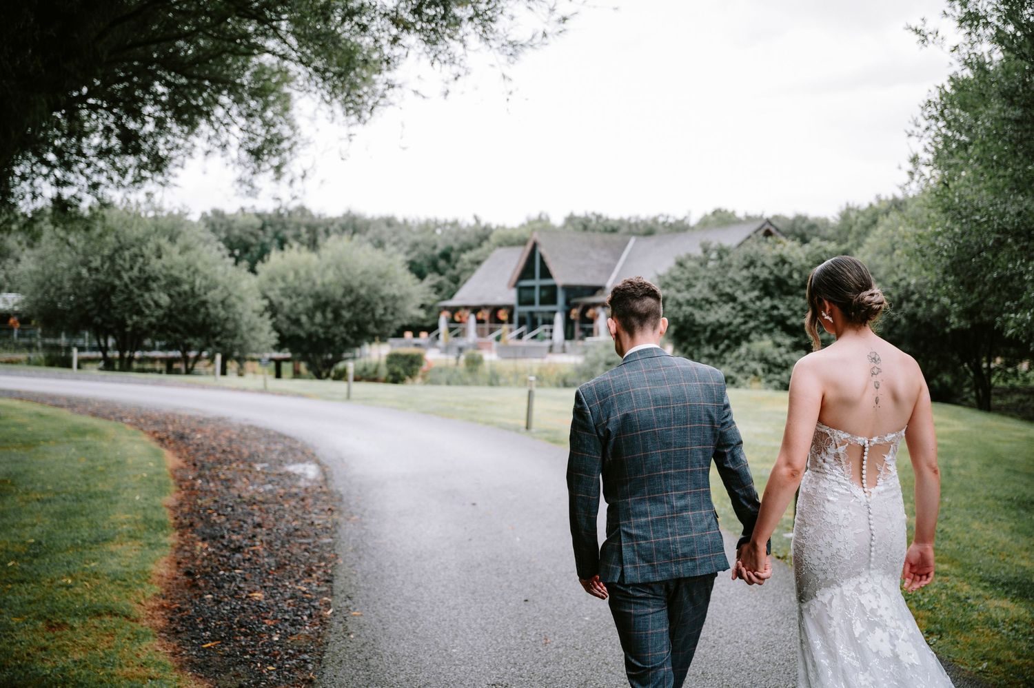 A couple walks down a winding path towards a rustic venue building on their wedding day.
