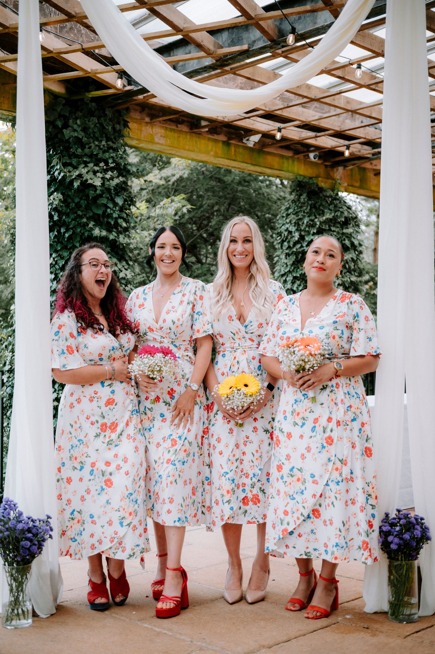 Bridesmaids in floral dresses and red shoes pose under decorated pergola.
