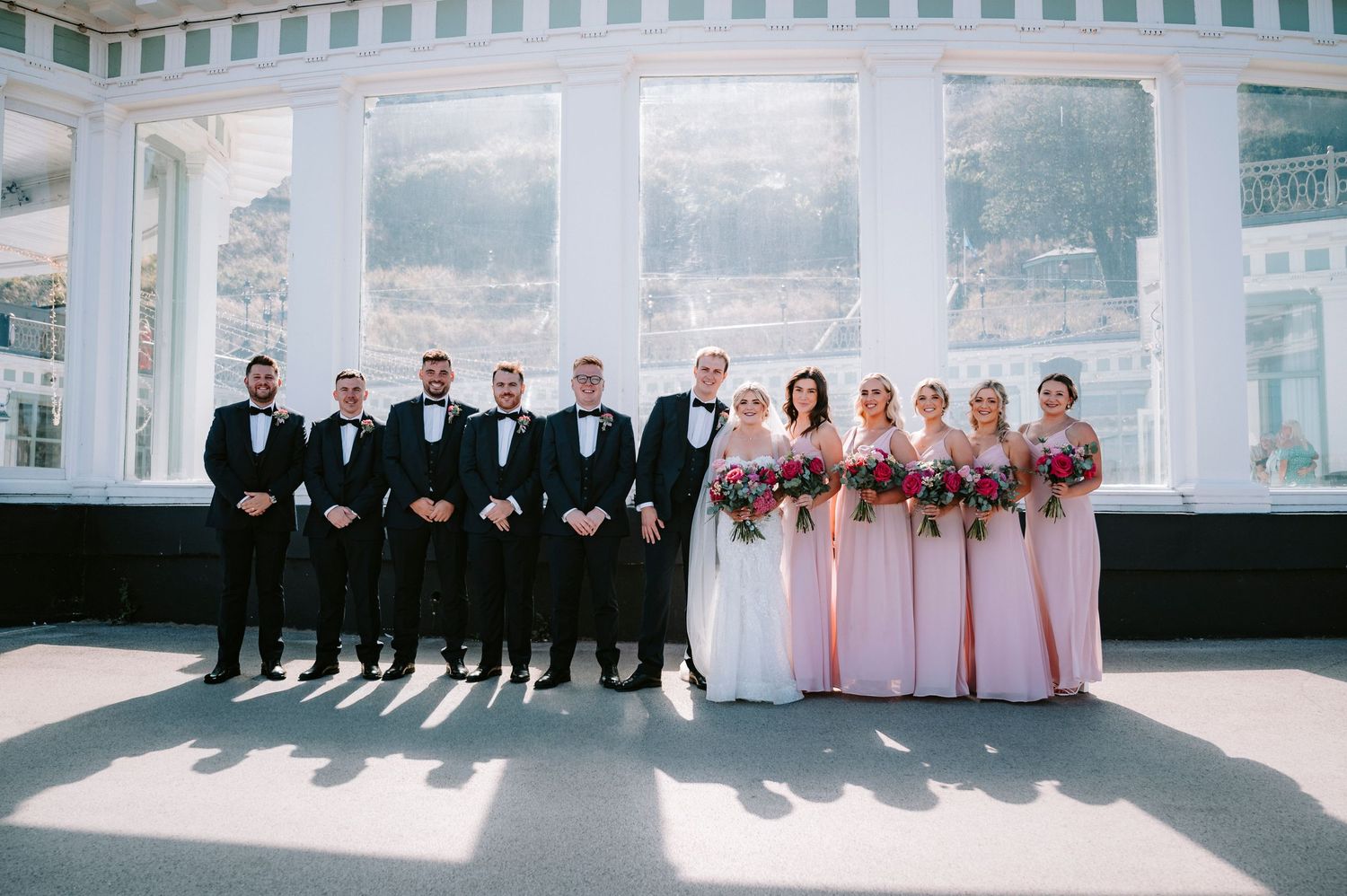 Wedding party lined up against white columns with bridesmaids in pink and groomsmen in black.