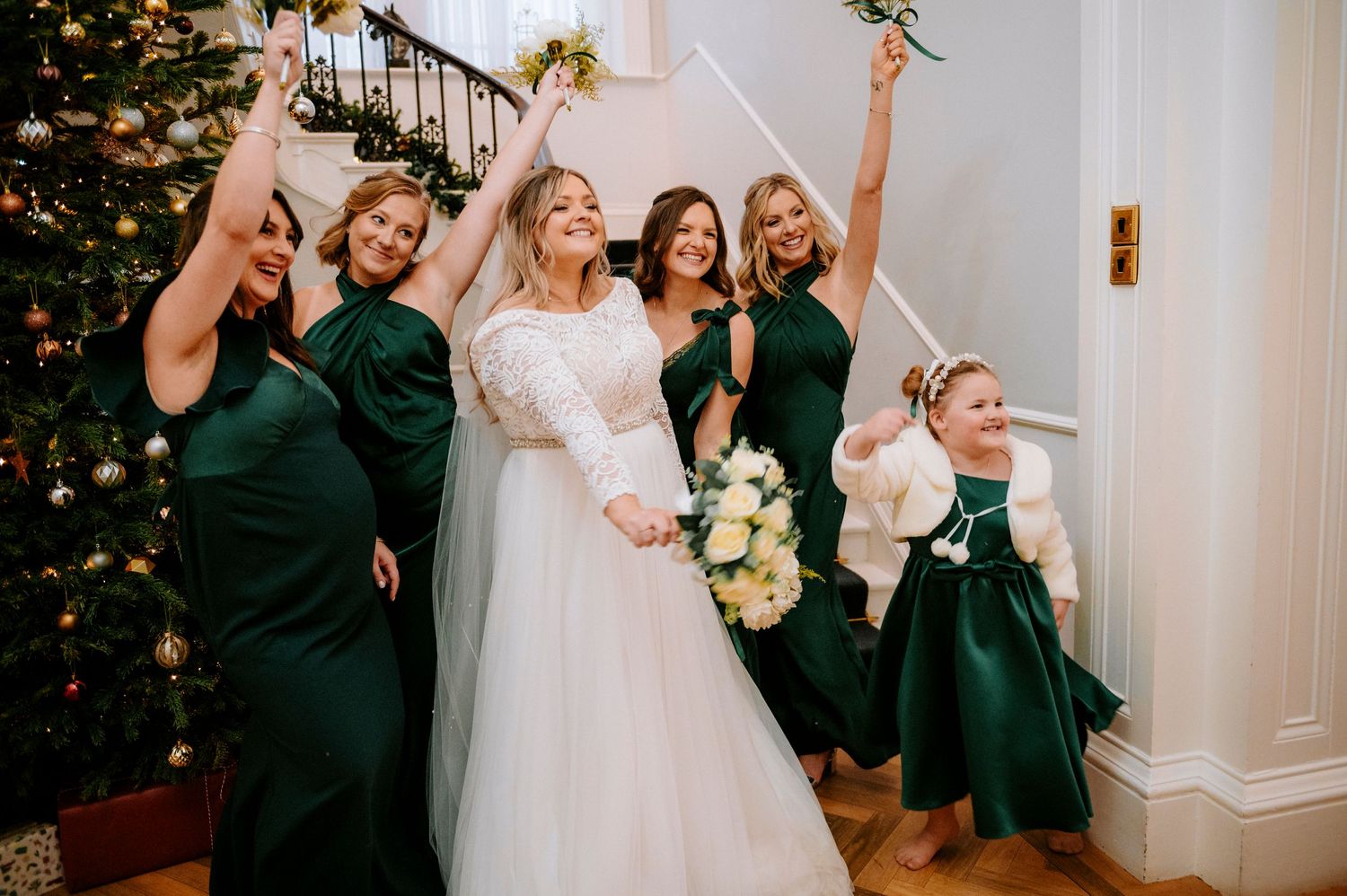 Wedding party celebrates joyfully in an elegant indoor venue with white decor and Christmas trees.