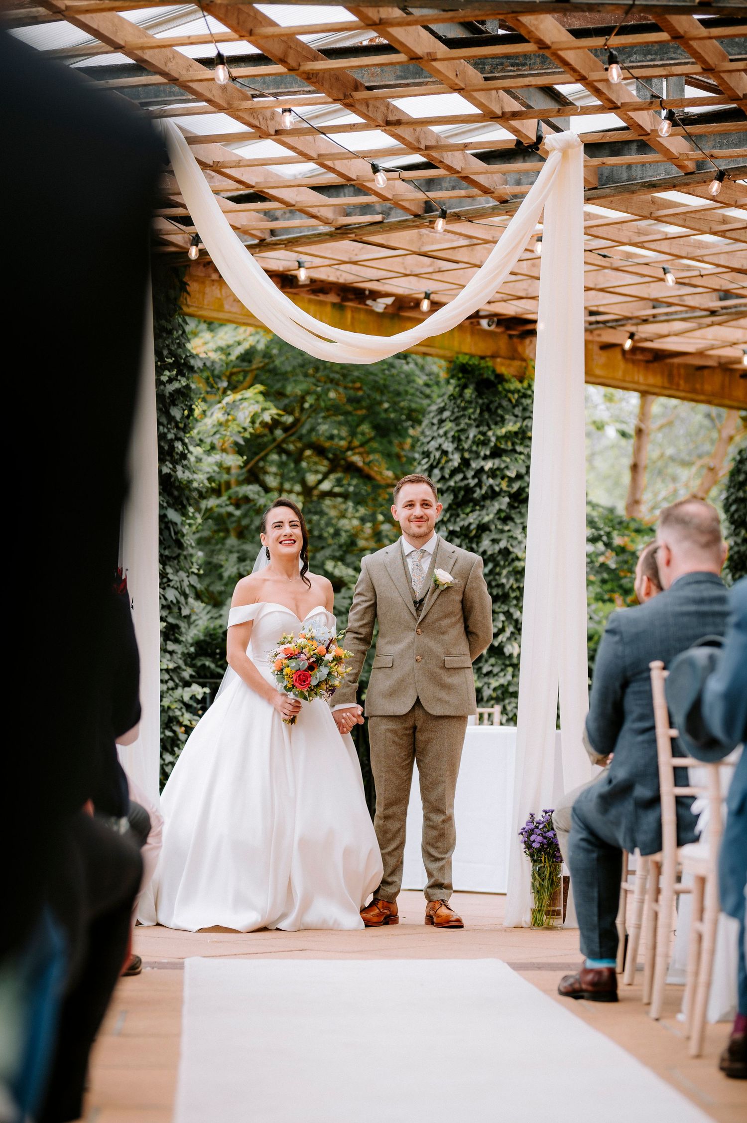 Wedding ceremony under wooden pergola decorated with white drapes and string lights.