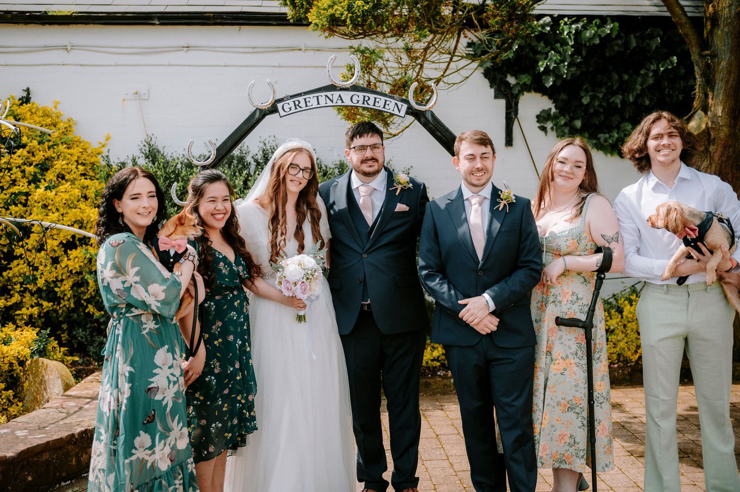 Wedding party poses together in formal attire with bridesmaids in sage green dresses at an outdoor venue.