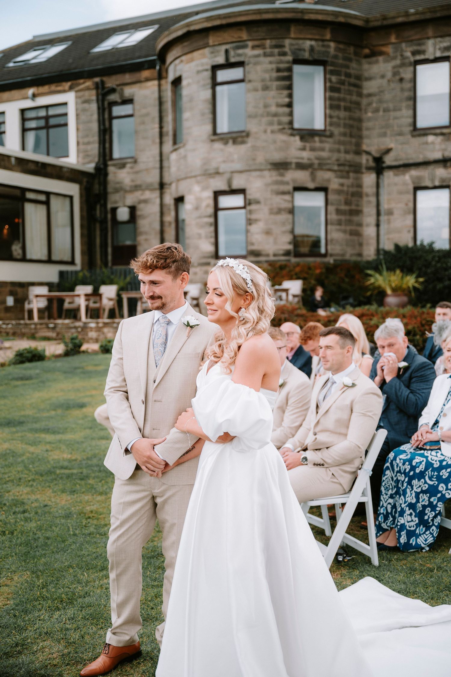 Wedding ceremony taking place outside a stone building with guests seated in white chairs.