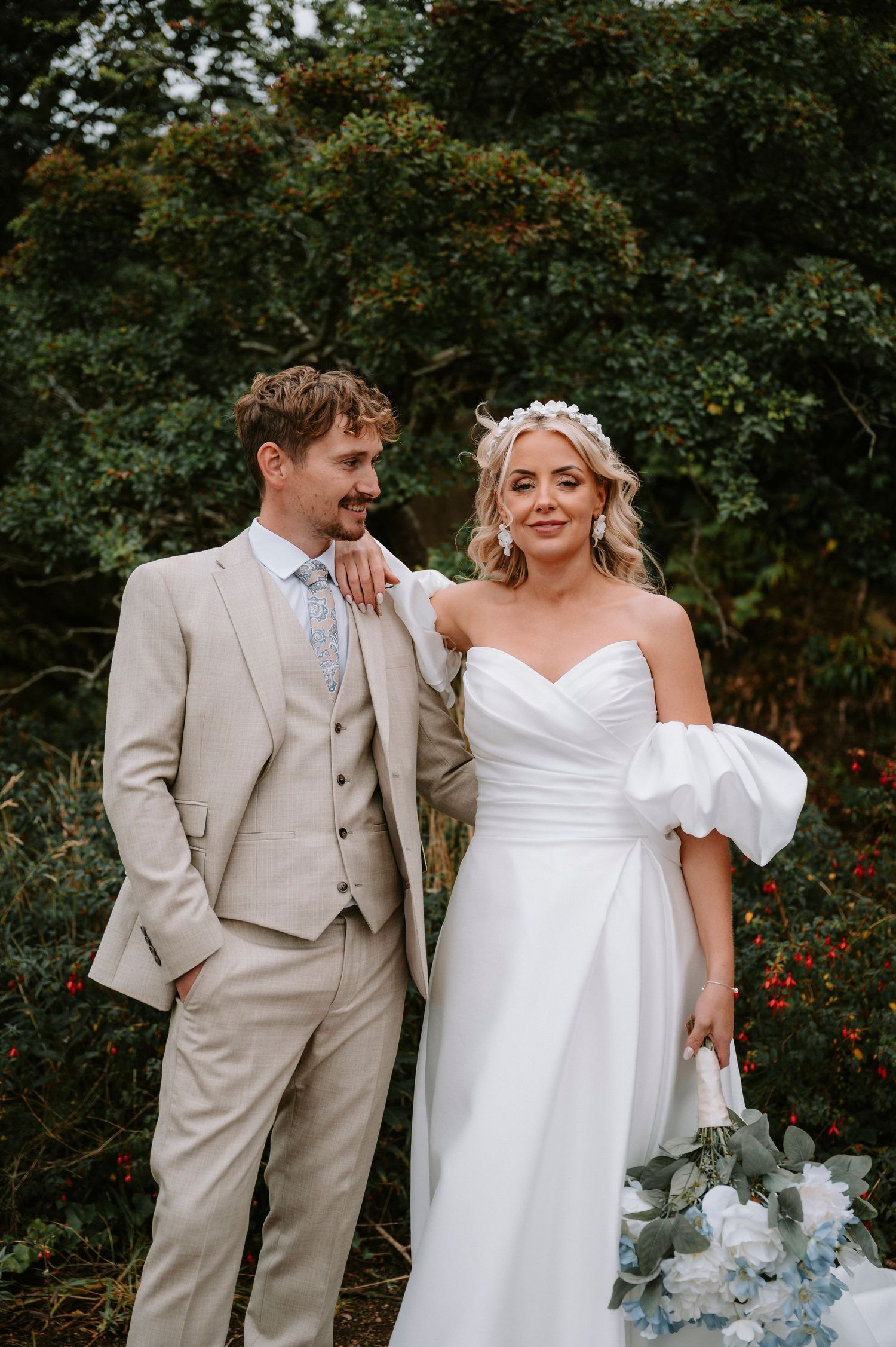 Newlyweds in beige and white attire pose together in garden setting with floral arrangements.