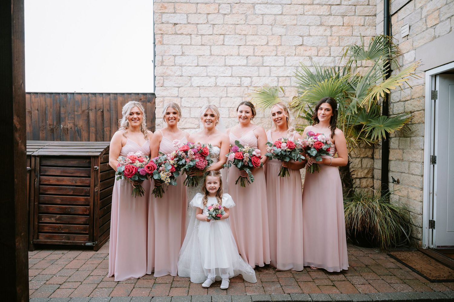 Wedding party lined up against white brick wall in pink dresses.