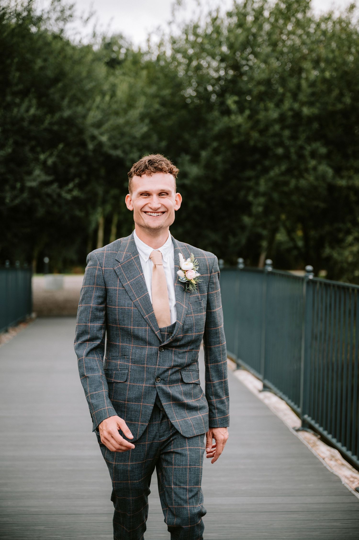 Groom wearing checkered grey suit with pink tie stands on wooden bridge.
