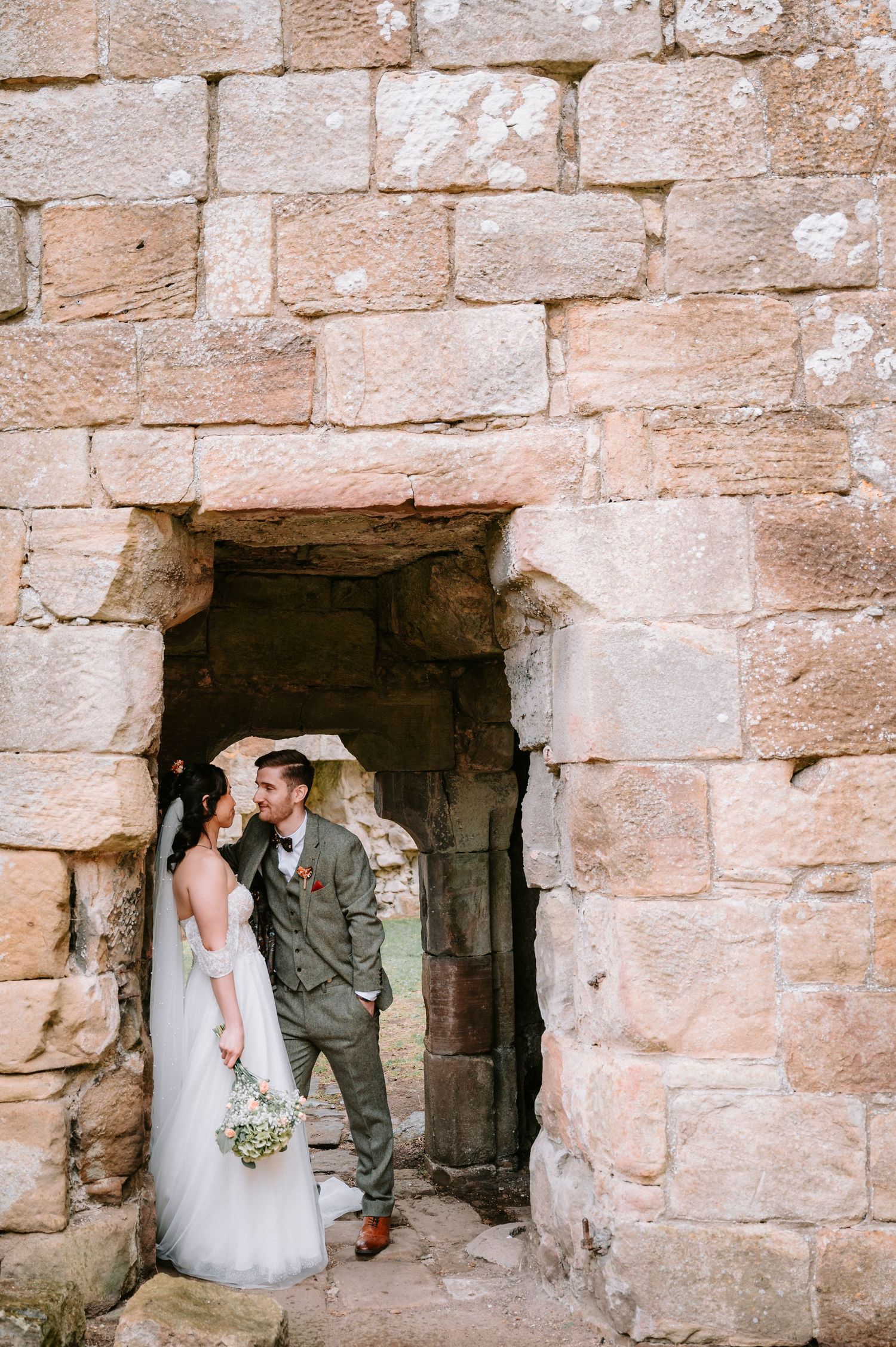 A romantic wedding photo of a couple standing in a rustic stone doorway archway with the bride holding a bouquet.