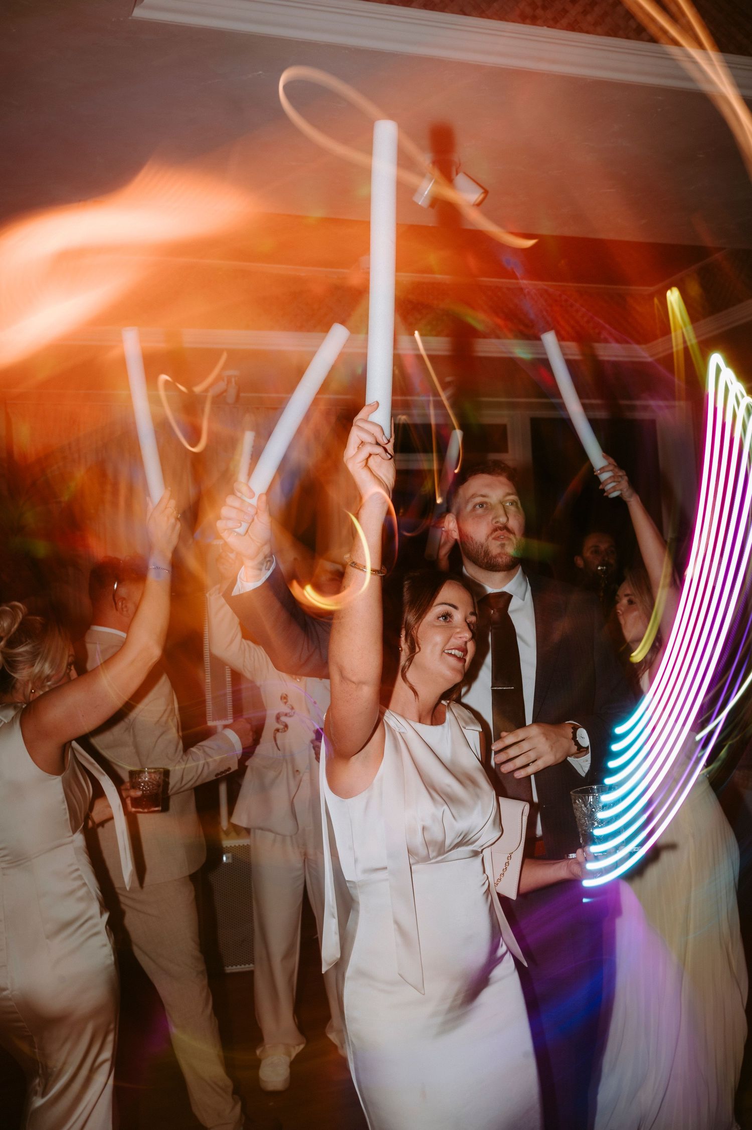 Party guests wave LED light sticks and glow sticks on a dark dance floor creating colorful light trails.