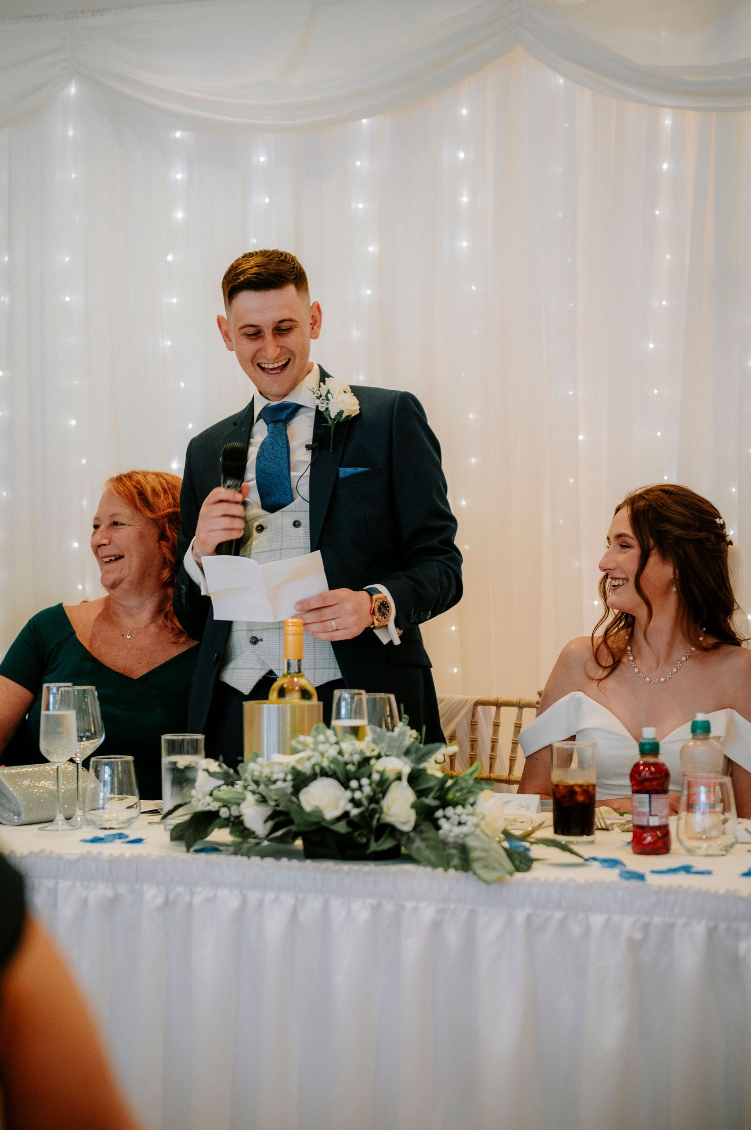Wedding reception scene showing guests and wedding party laughing at head table with floral decorations.