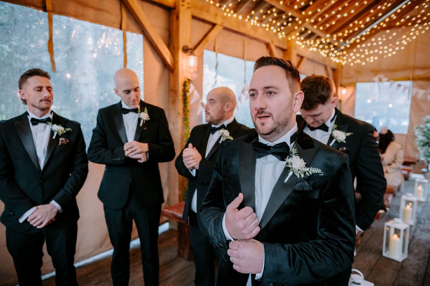 Groomsmen in black tuxedos gather before the ceremony in a rustic venue with string lights.