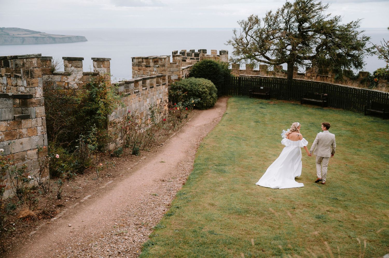 Stone castle wall pathway leading to wedding couple on grassy grounds.