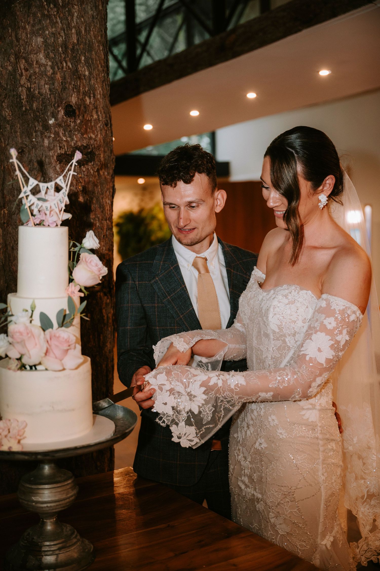 Newlyweds cut their elegant white wedding cake decorated with delicate flowers in a rustic indoor venue.