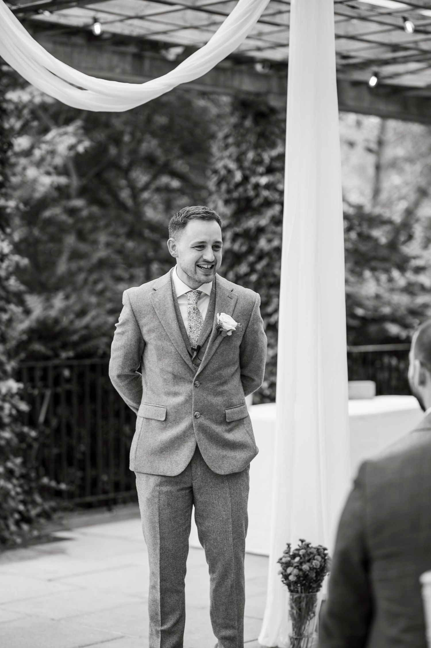 Groom in gray suit laughs joyfully during outdoor ceremony.