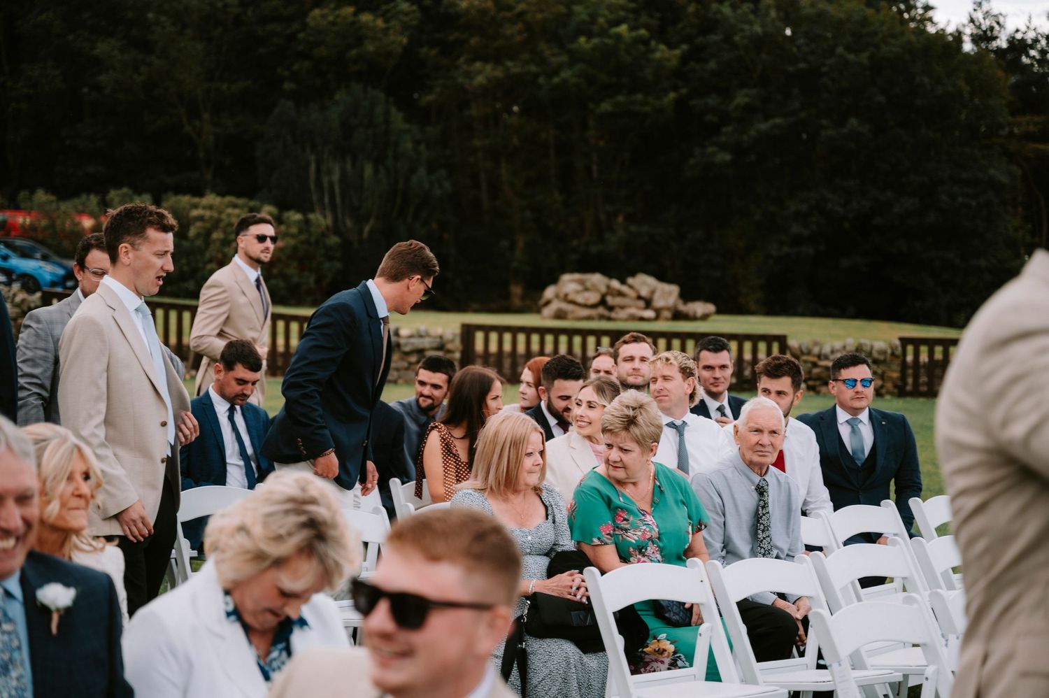 Wedding guests in formal attire gathered outdoors for the ceremony on a sunny day.