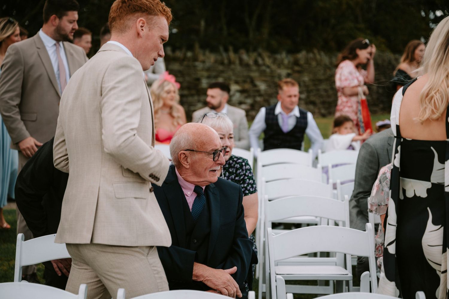 Wedding guests in formal attire gathered around white chairs at outdoor ceremony.