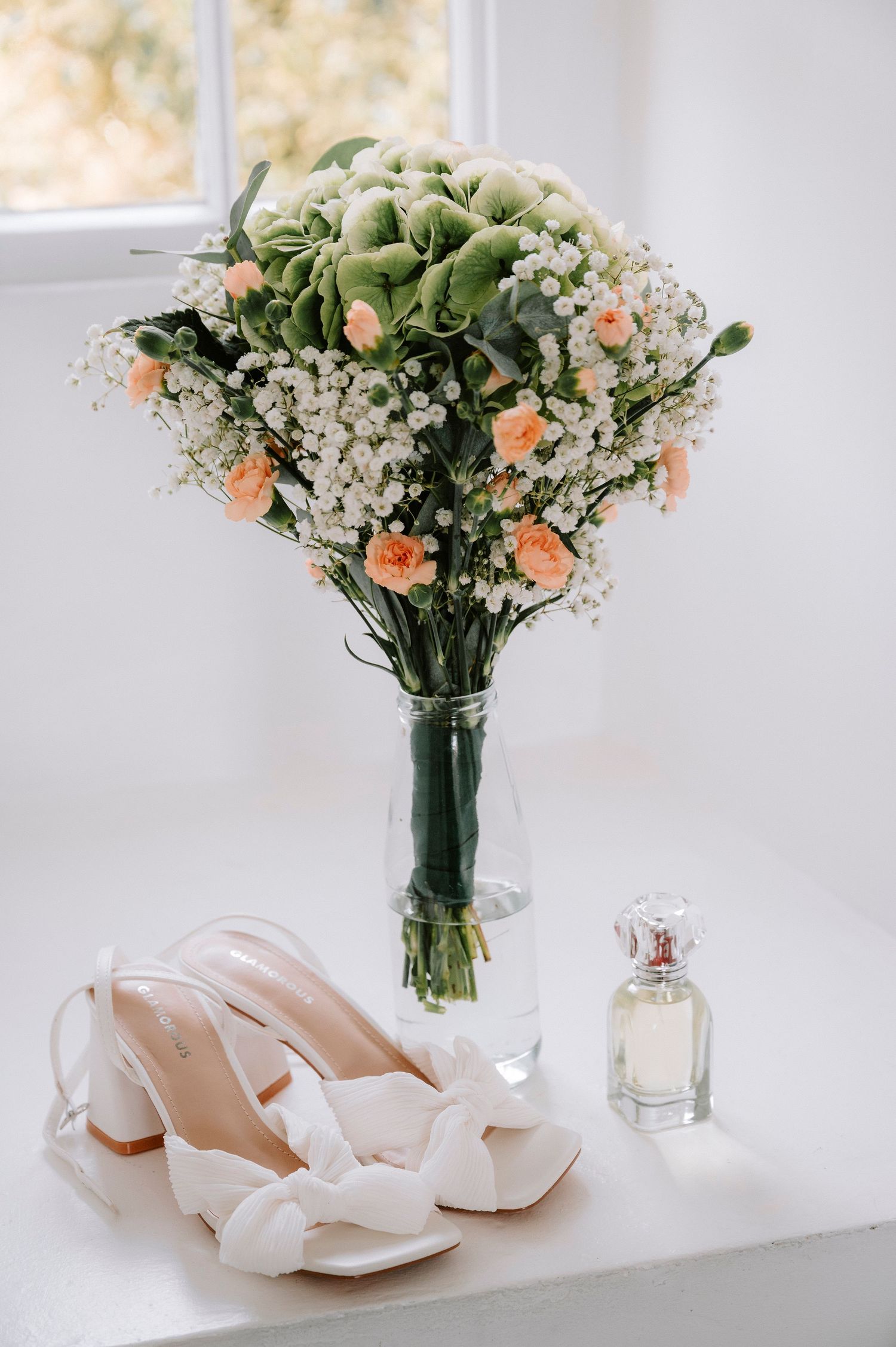 White bridal shoes displayed next to a delicate floral bouquet with orange and white flowers in a glass vase.