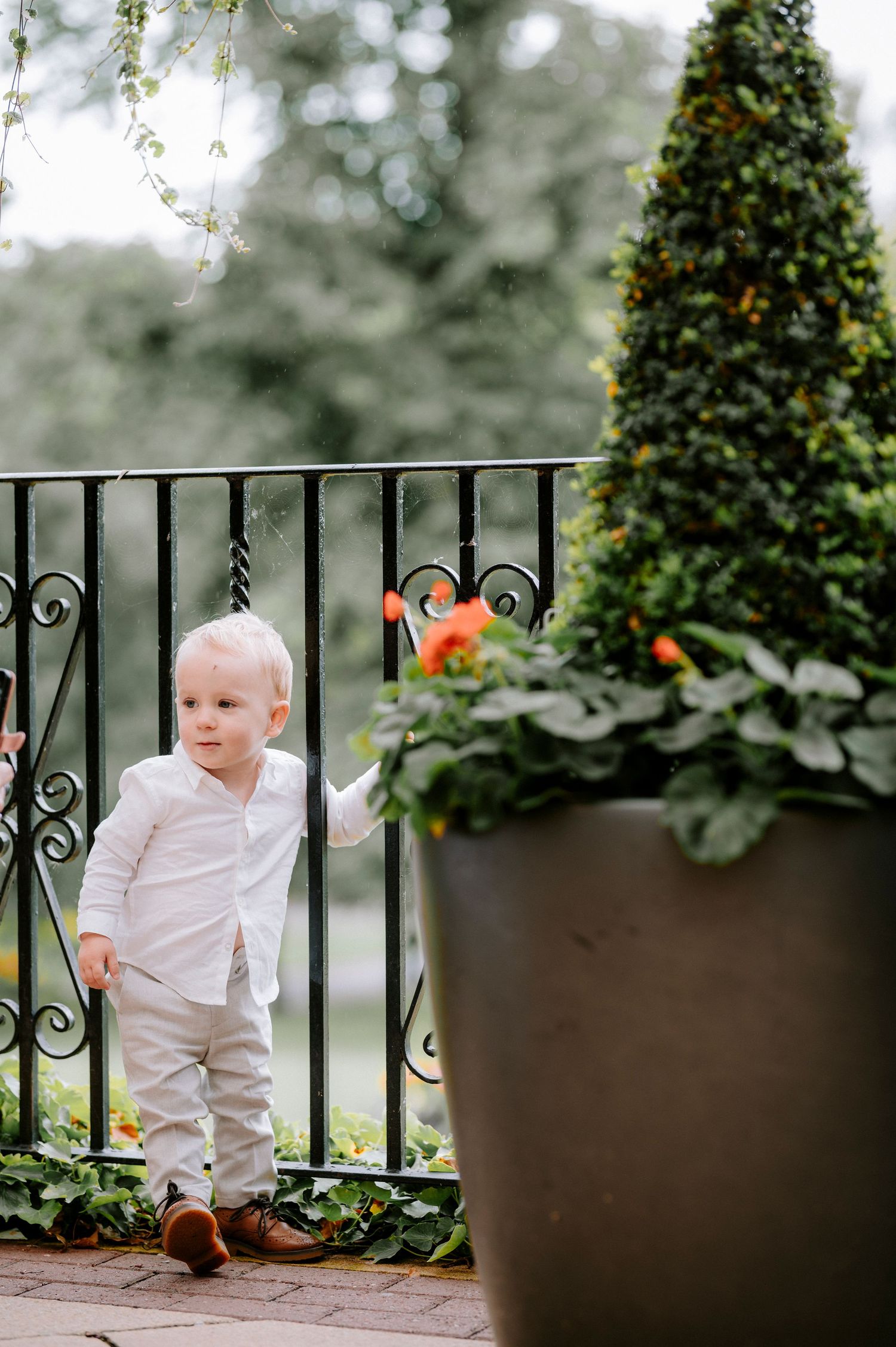 Small child in white outfit plays near decorative potted plants and railing.