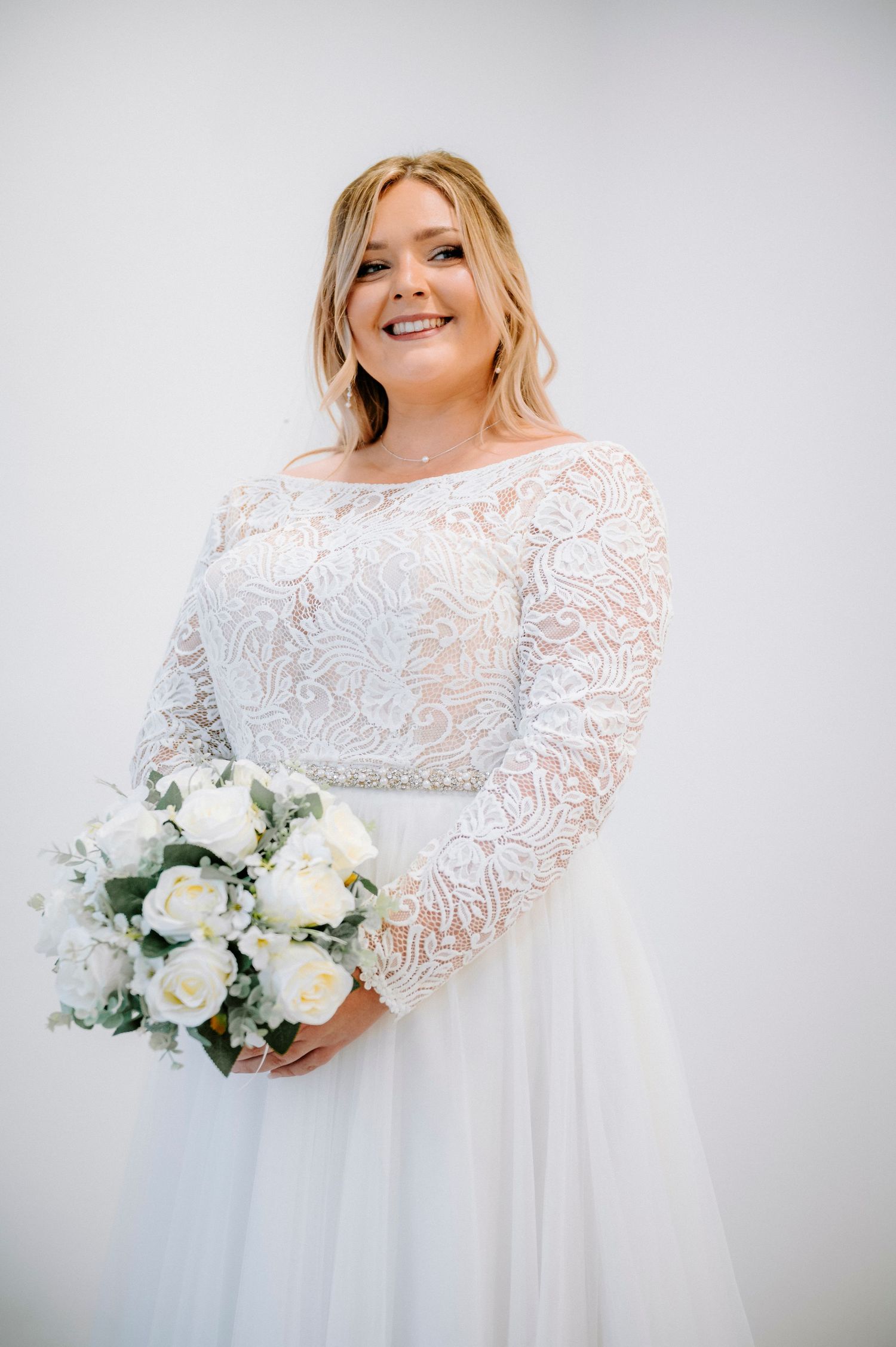 A radiant bride poses in a long-sleeved lace wedding gown while holding a white floral bouquet.
