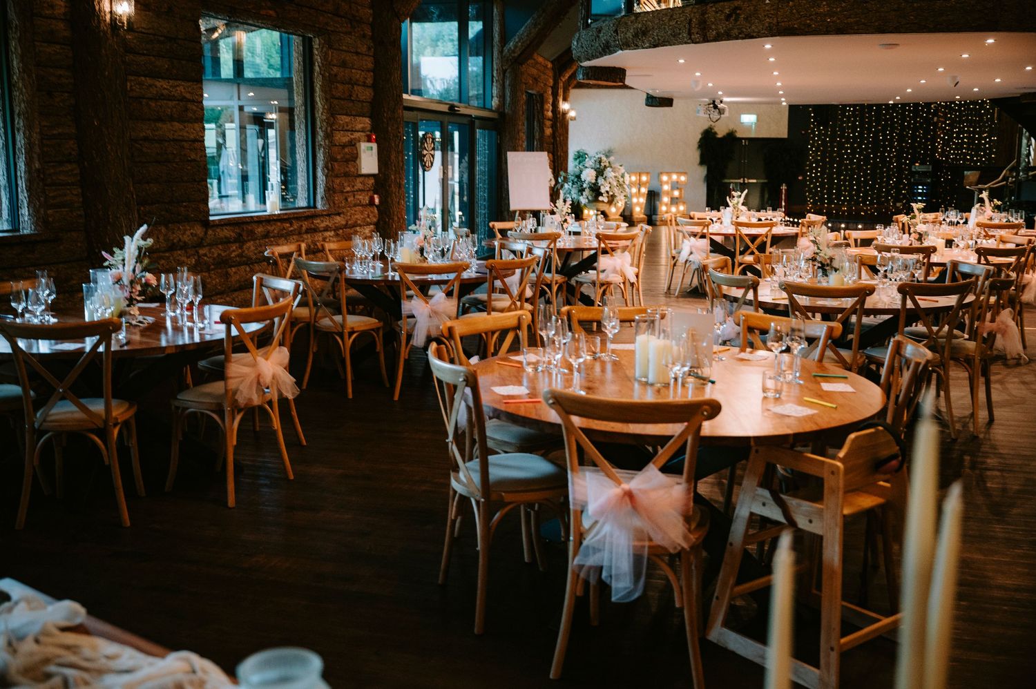 Rustic wedding reception venue with wooden tables and chairs set for dinner under warm lighting.