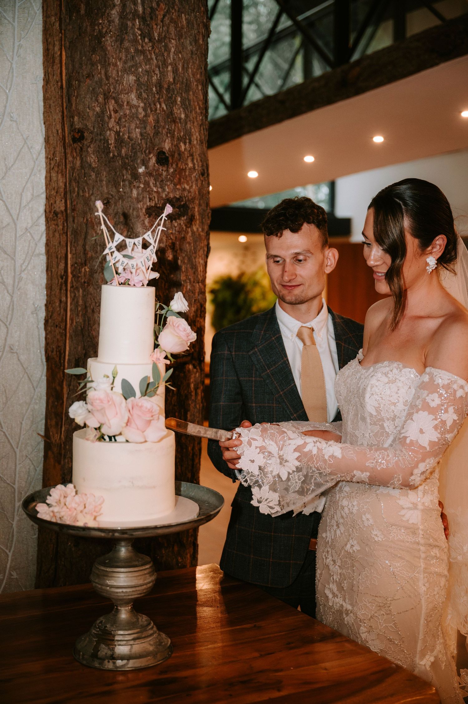 Elegant white wedding cake with delicate floral details being cut by newlyweds at an indoor reception.